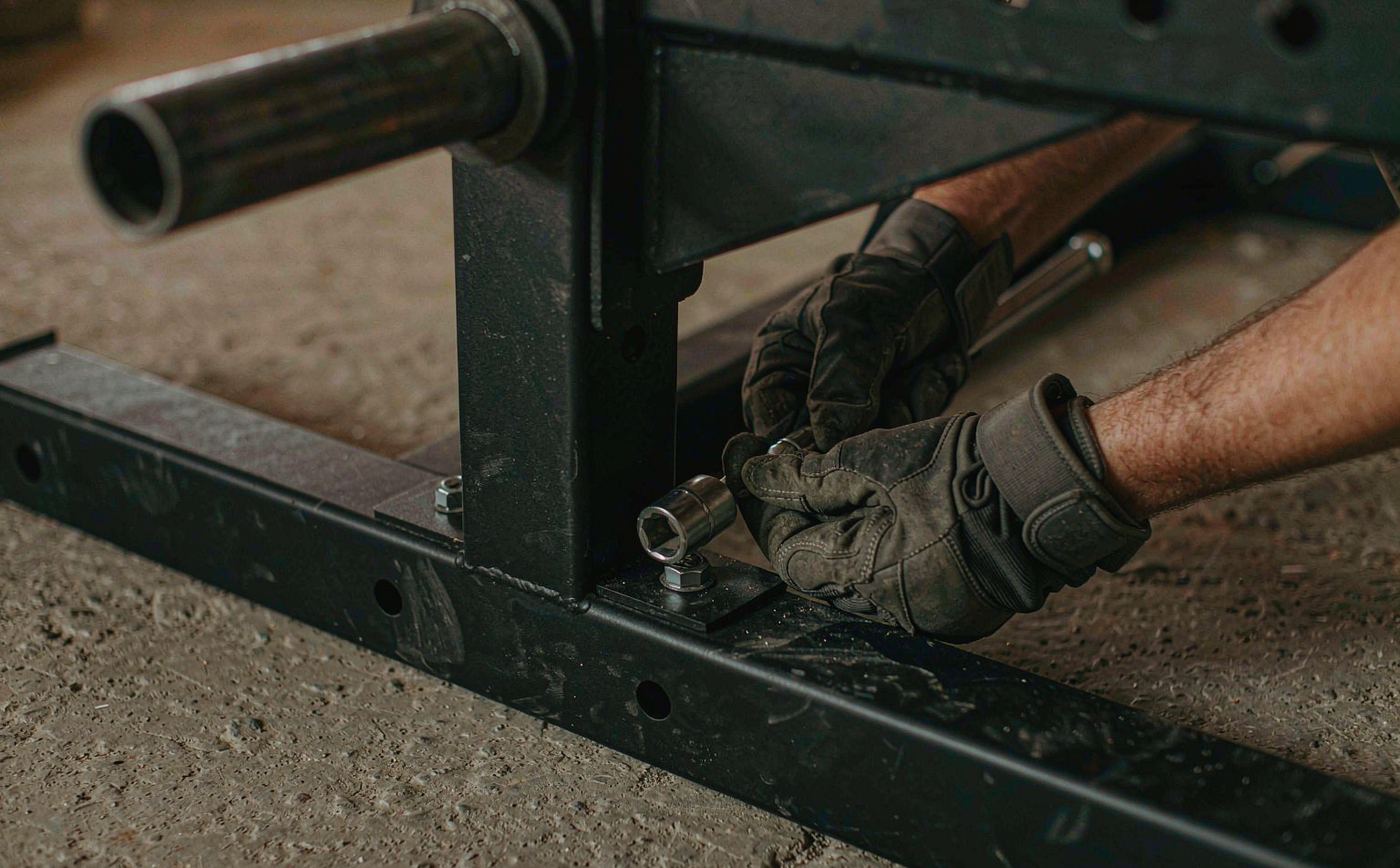 A gloved hand tightening the base bolt of a black steel power rack with a socket wrench on a concrete floor.
