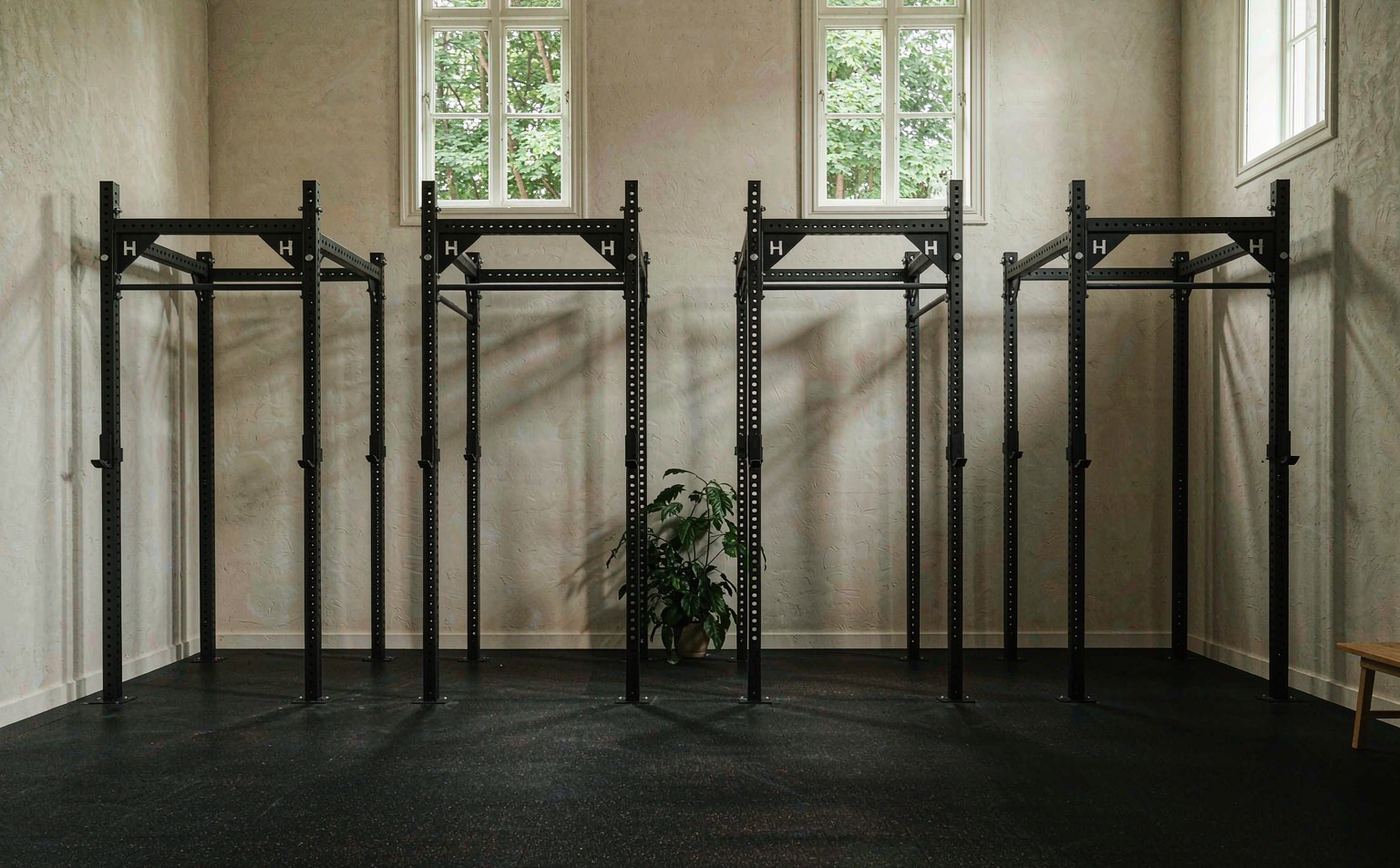 Four black power racks in a row against a white plaster wall in a small strength studio with polished concrete floors.