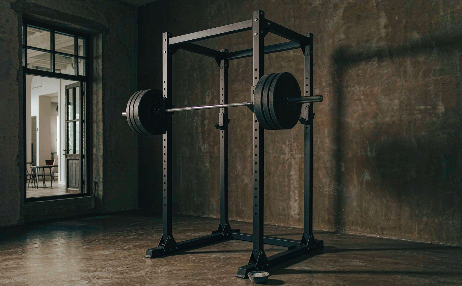 A matte black Xiaole power rack with a loaded barbell standing on polished concrete in a garage gym, lit by a tall window.